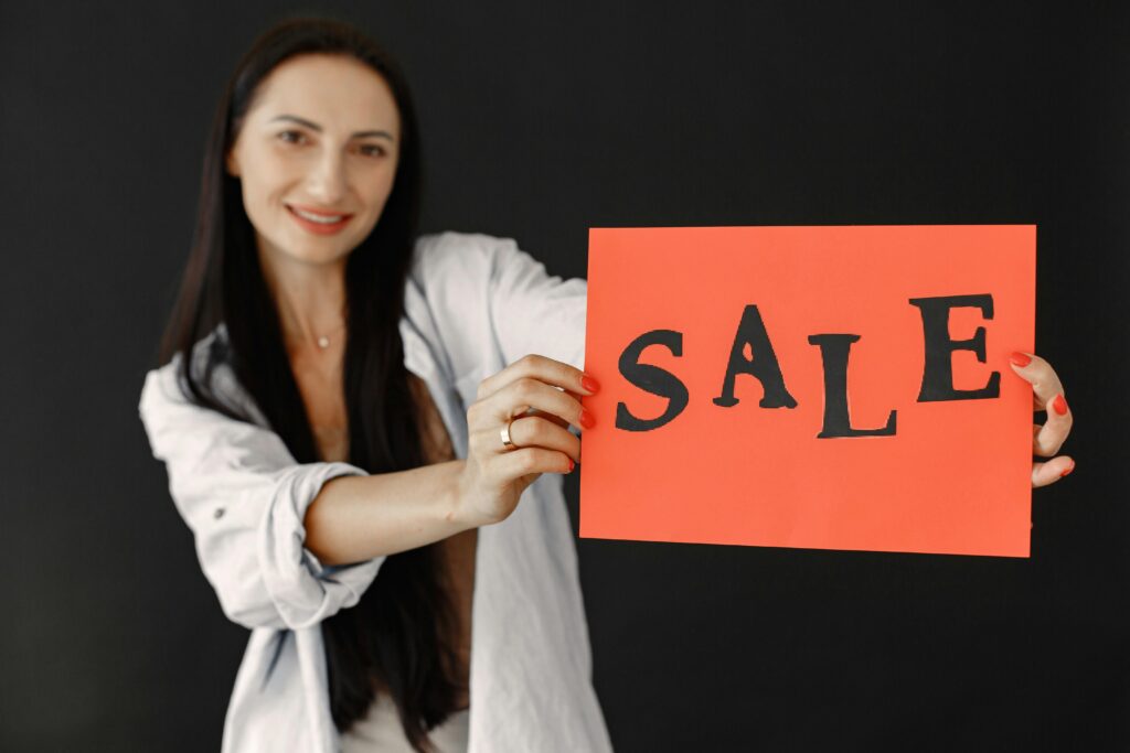 Smiling woman with long black hair holds a vibrant sale sign against a dark background.